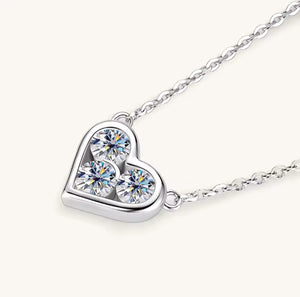 A silver heart-shaped pendant necklace featuring three round-cut clear crystals, shown on a delicate silver chain against a light cream background.