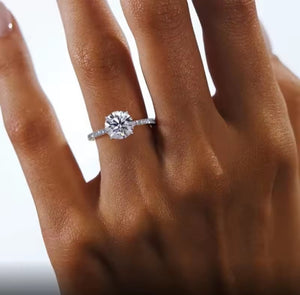 A close-up of a hand wearing a silver engagement ring with a round-cut clear center stone and small side stones, set against a bright white background.