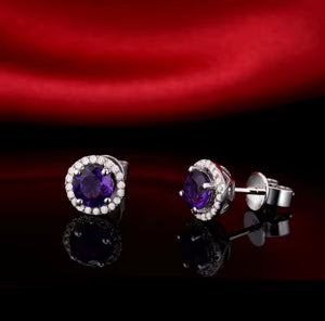 A pair of oval halo stud earrings with deep purple natural amethyst center stones and clear accent natural diamond stones in a silver-tone setting, displayed on a glossy black surface with a red satin fabric backdrop.