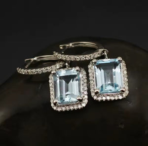 A pair of white gold earrings with rectangular light blue topaz gemstone centres diamond halo accents, displayed on a dark reflective surface.