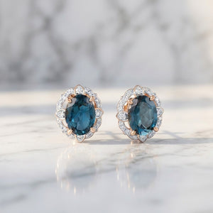 A pair of oval stud earrings with deep blue topaz gemstones surrounded by small clear natural diamond stones in a rose gold setting, displayed on a light marble surface.