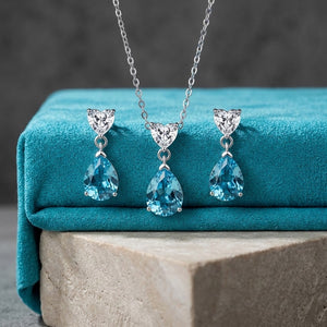 Silver-tone necklace and matching drop earrings with heart-shaped clear crystal tops and pear-shaped blue gemstones, displayed on a teal velvet jewelry box.
