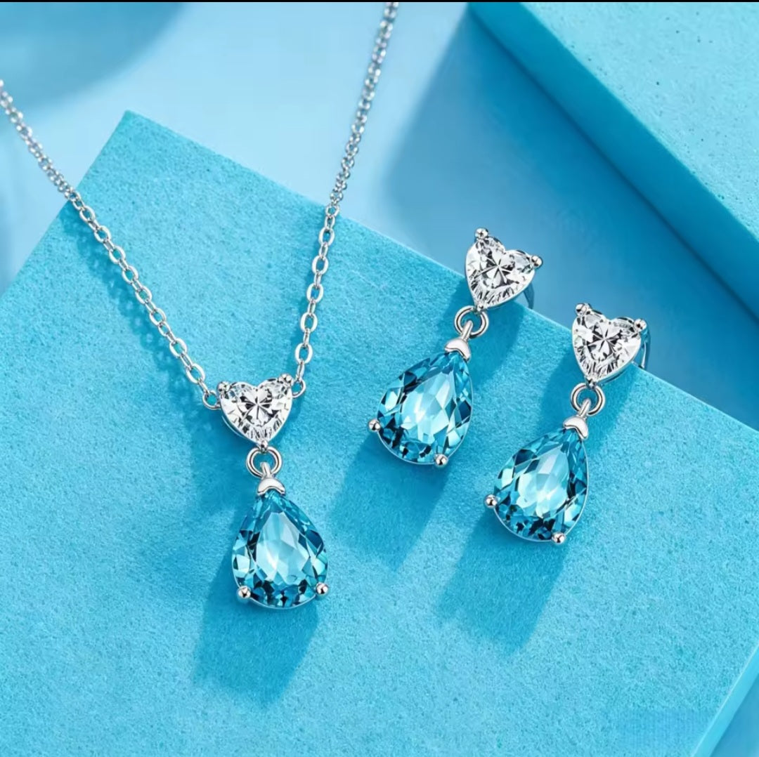 Silver jewellery set featuring a necklace and matching drop earrings with clear heart-shaped crystals and light blue teardrop-shaped crystals, displayed on a textured light blue surface.