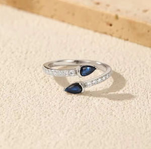 A silver wrap-style ring featuring two teardrop-shaped dark blue gemstones and rows of small clear stones along the band, photographed on a light beige textured surface.