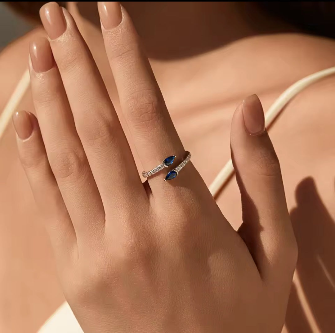 A close-up of a hand wearing a silver ring with two marquise-cut blue gemstones and small clear stones along the band, set against a warm-toned skin and neutral background.
