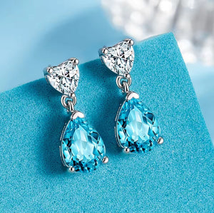 A pair of silver drop earrings with heart-shaped clear gemstones and large faceted light blue teardrop gemstones, displayed on a textured light blue surface.