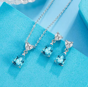 A jewellery set featuring a silver necklace and matching drop earrings with light blue teardrop-shaped crystals and clear triangular crystals, displayed on a textured light blue surface.