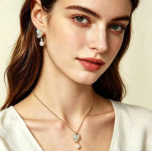 Close-up of a woman wearing a delicate gold necklace with a butterfly-shaped pendant featuring iridescent blue-green stones and white pearls, along with a matching drop earring, against a soft beige background.