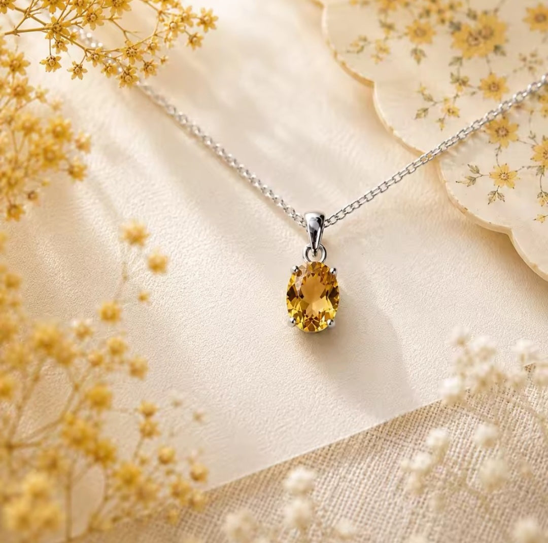 A silver chain necklace with a teardrop-shaped yellow natural citrine gemstone pendant, displayed on a cream-colored fabric with delicate dried flowers.