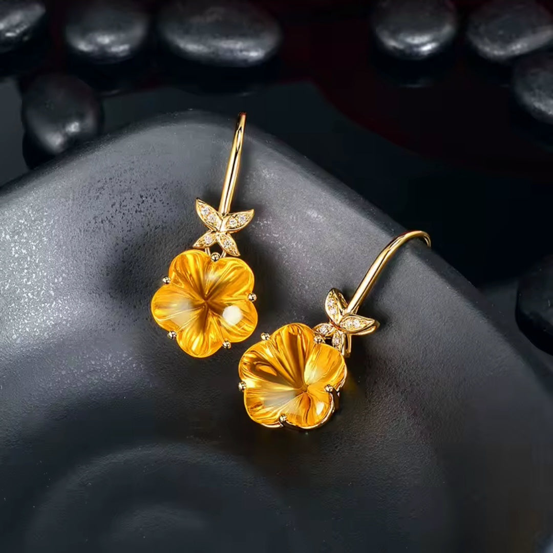 A pair of gold flower drop earrings with yellow-orange natural citrine, translucent petals and small clear natural diamond stones in the leaf settings, displayed on a dark textured surface with smooth dark stones in the background.