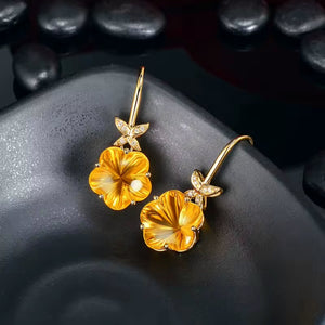 A pair of gold flower drop earrings with yellow-orange natural citrine, translucent petals and small clear natural diamond stones in the leaf settings, displayed on a dark textured surface with smooth dark stones in the background.