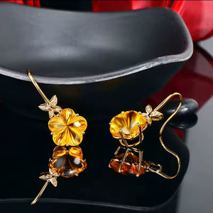 A pair of gold drop earrings with large yellow-orange citrine flower charms and small leaf details with natural diamonds, displayed on a black stand with a reflective surface.