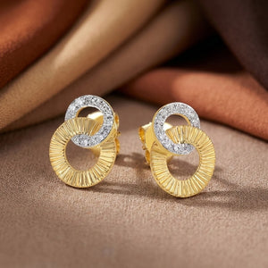 A pair of gold hoop earrings with a textured, sunburst-style gold lower half and a yellow gold upper half paved with small diamond stones, displayed on a textured brown fabric background.