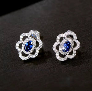 A pair of white gold stud earrings, each featuring a central oval blue sapphire stone surrounded by a halo of small clear natural diamond stones, displayed on a dark textured surface.