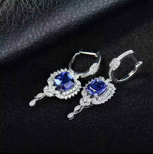 A pair of white gold earrings with large square blue gemstones surrounded by small clear stones, displayed on a textured black surface.