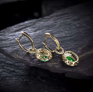 A pair of gold hoop drop earrings with filigree detailing and oval green emeralds and natural diamonds, displayed on a dark textured slate surface.