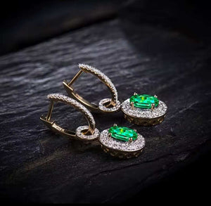 A pair of gold drop hoop earrings with oval green emerald gemstones and surrounding diamond stones, displayed on a dark textured surface.