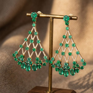A pair of gold-tone dangle earrings with green natural gemstones and diamonds, displayed on a gold stand against a warm beige textured background.