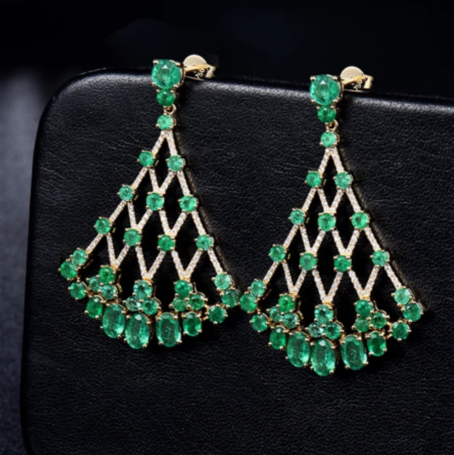 A pair of emerald and diamond drop earrings, with a solid 18K yellow gold band, resting on a black surface.