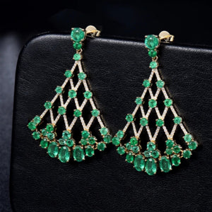 A pair of emerald and diamond drop earrings, with a solid 18K yellow gold band, resting on a black surface.
