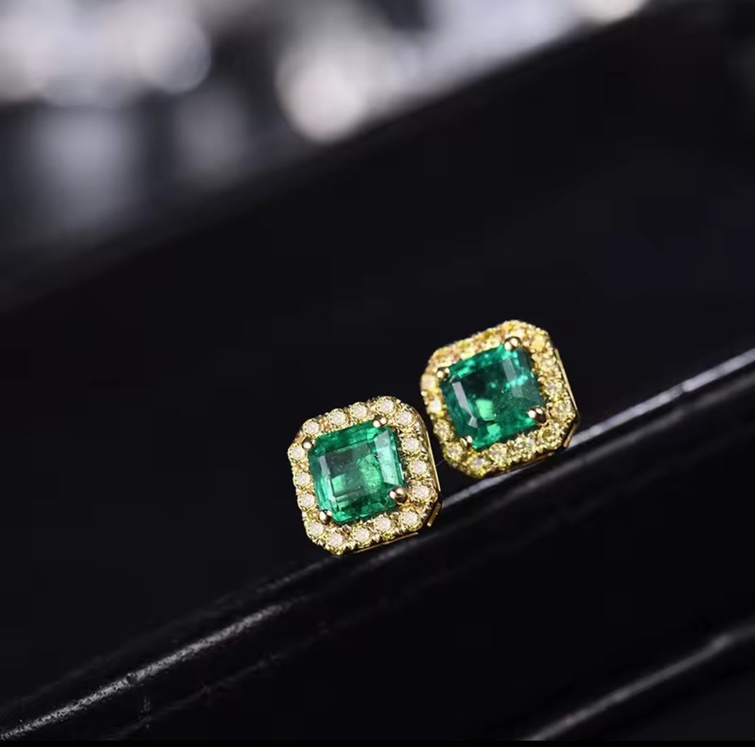 A pair of gold stud earrings with square-cut green emerald-cut center stones surrounded by small clear pavé stones, displayed on a black surface.