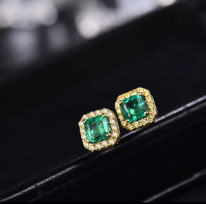 A pair of gold stud earrings with square-cut green emerald-cut center stones surrounded by small clear pavé stones, displayed on a black surface.