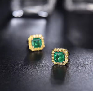 A pair of gold square stud earrings with emerald-cut green gemstones and small clear accent stones, displayed on a dark textured surface.