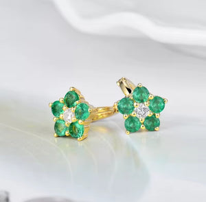 A pair of gold flower-shaped stud earrings with six green emerald-cut stones each and a small clear center stone, shown on a soft white surface.