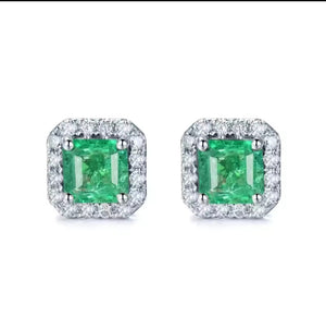 A pair of square halo stud earrings featuring vibrant green emerald-cut center stones surrounded by a border of small clear stones in a silver-toned setting, shown on a plain white background.