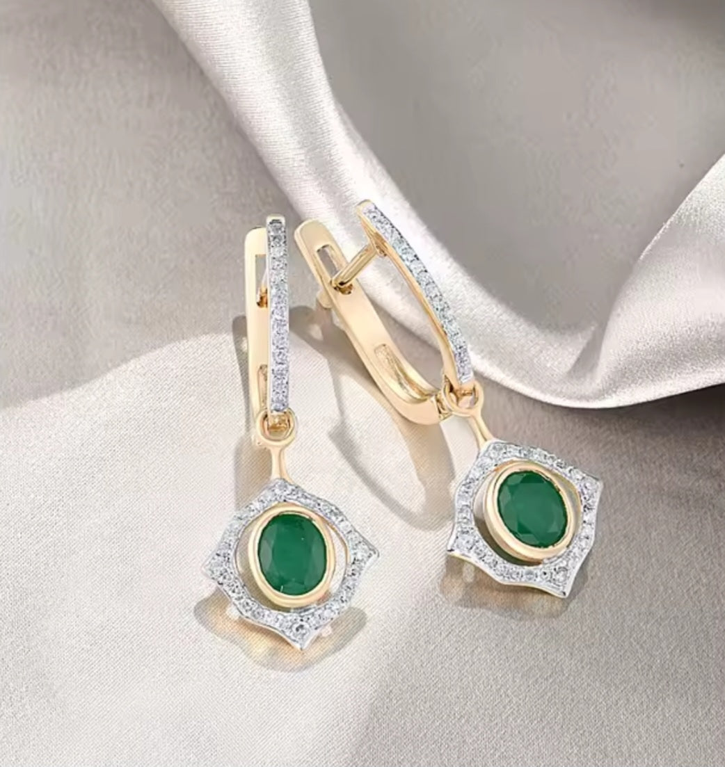 A pair of gold and silver drop earrings with oval green emerald gemstones, accented by clear natural diamonds, displayed on a draped champagne satin fabric.