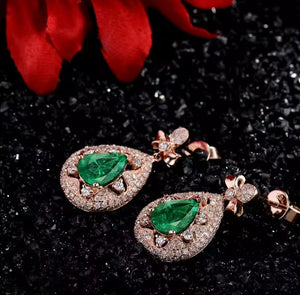 A pair of rose gold teardrop-shaped drop earrings with large green emerald-cut center stones and pavé-set clear accent stones, displayed on a dark glittery surface with red flower petals in the background.