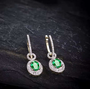 A pair of gold drop hoop earrings with oval green natural emerald gemstones and halo diamond accents, displayed on a dark textured stone surface.