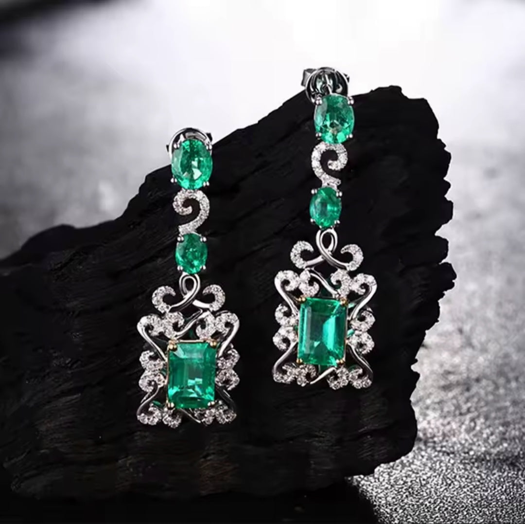 A pair of white gold dangle earrings with large emerald-cut green gemstones and smaller round green stones, accented with clear stones, displayed on a dark textured stone surface.