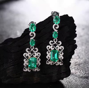 A pair of white gold dangle earrings with large emerald-cut green gemstones and smaller round green stones, accented with clear stones, displayed on a dark textured stone surface.