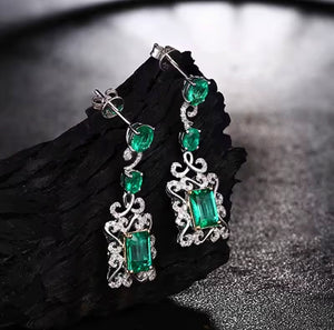 A pair of white gold drop earrings with large green emerald-cut centre stones and smaller green natural emerald stones, accented by clear stones, displayed on a dark textured surface.