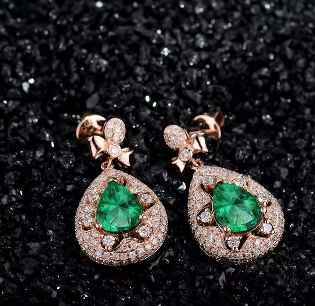 A pair of rose gold drop earrings featuring large green emerald-cut center stones surrounded by small clear pavé stones, displayed on a textured black background.