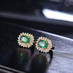 A pair of gold oval stud earrings featuring green emerald-cut centre stones surrounded by clear baguette and round diamonds, displayed on a dark reflective surface.