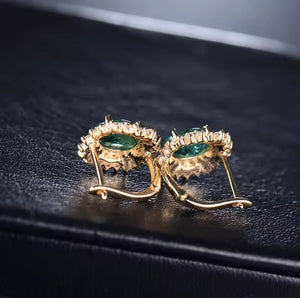A pair of gold hoop earrings with oval green emerald gemstones and small clear natural diamond stones around the edges, displayed on a textured black surface.