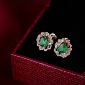 A pair of rose gold floral stud earrings with pear-cut green emerald and small clear natural diamond stones, displayed on a red textured jewellery box.