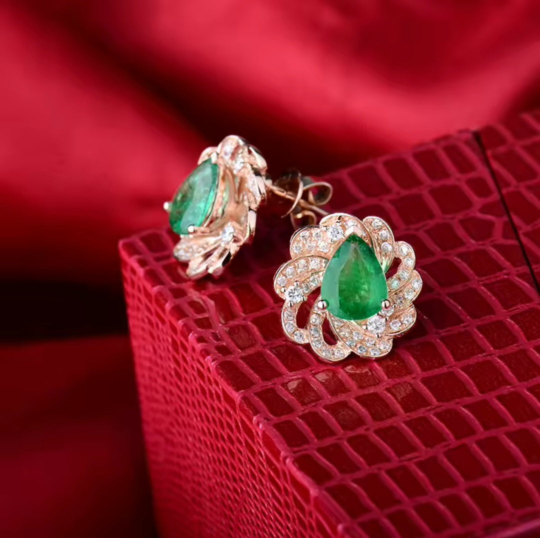 A pair of rose gold statement earrings with large green emerald gemstones and pave-set clear natural diamond stones in a floral scroll design, displayed on a textured red box against a red satin background.
