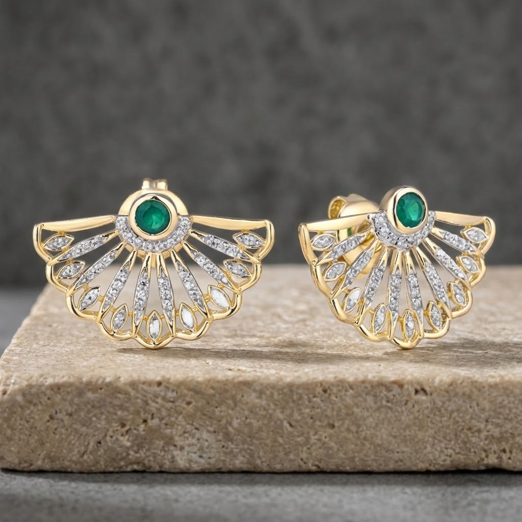 A pair of gold fan-shaped stud earrings with green natural emerald oval center stones and rows of clear pavé natural diamond stones, displayed on a stone slab against a dark gray background.