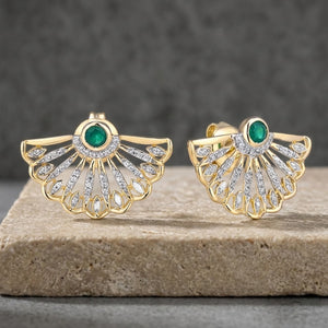 A pair of gold fan-shaped stud earrings with green natural emerald oval center stones and rows of clear pavé natural diamond stones, displayed on a stone slab against a dark gray background.