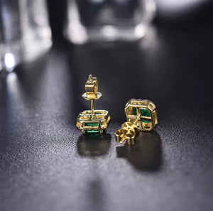 A pair of gold geometric stud earrings with rectangular green emerald-cut stones, shown from both front and side angles on a dark reflective surface.