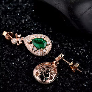 Rose gold drop earrings displayed on a textured black background. One earring features a marquise-cut green gemstone surrounded by small clear stones, and the other features an intricate filigree heart-shaped drop.