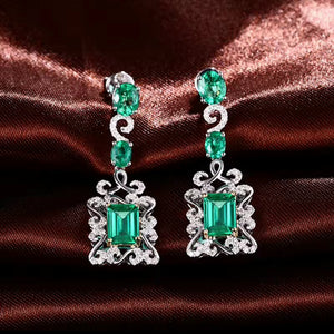 A pair of white gold drop earrings with natural emerald green gemstones and small clear natural diamond accent stones, displayed on a dark brown satin fabric background.
