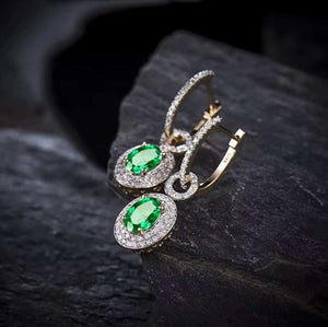 A pair of gold drop hoop earrings with oval green emeralds gemstones surrounded by small diamond stones, displayed on a dark textured stone surface.