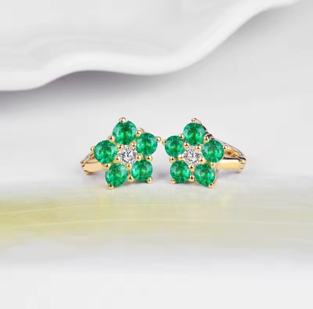 A pair of gold flower-shaped rings with five green emerald-cut stones on each ring and a small clear center stone, displayed on a soft white surface.