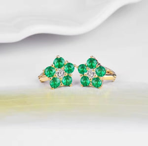 A pair of gold flower-shaped rings with five green emerald-cut stones on each ring and a small clear center stone, displayed on a soft white surface.