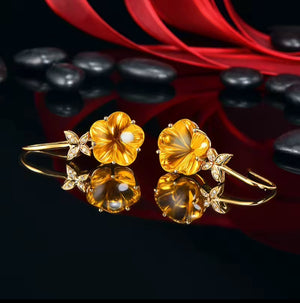 A pair of gold natural citrine flower-shaped earrings with natural diamonds and small leaf details, displayed on a glossy black surface with red fabric and dark stones in the background.