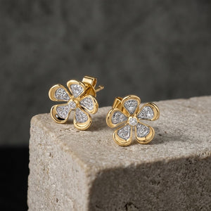 A pair of gold flower-shaped stud earrings set with clear natural diamonds stones, displayed on a textured stone block against a dark grey background.